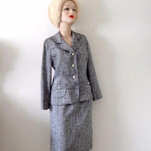 Puede incluir: Un traje de falda de tweed gris y blanco con una camisa blanca abotonada y un sombrero de piel blanca. El traje tiene una chaqueta entallada con cuello y bolsillos. La falda es de largo hasta la rodilla y tiene una silueta recta.