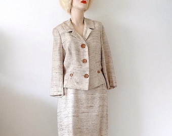 May include: A beige skirt suit with a matching jacket and skirt. The jacket has a notched collar, long sleeves, and four brown buttons down the front. The skirt is knee-length and has a straight silhouette.