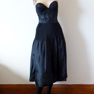 May include: A black lace bustier and a black skirt with pleats. The skirt is knee-length and has a fitted waist. The mannequin is wearing black shoes.