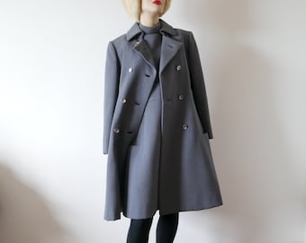 May include: A gray double-breasted coat and matching dress on a mannequin. The coat has large buttons and a flared skirt. The mannequin wears black tights and black pointed-toe shoes. A light-colored fur hat completes the ensemble.