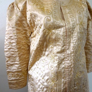 Peut inclure: Veste en satin cr&egrave;me avec broderies dor&eacute;es et petites perles dor&eacute;es. La veste a un col rond et des manches longues avec des &eacute;paules bouffantes. La broderie forme des motifs floraux et tourbillonnants.