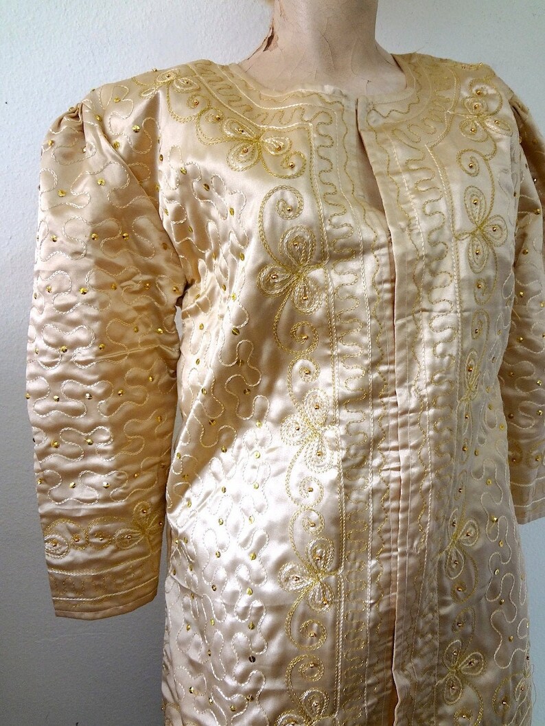 Peut inclure: Veste en satin cr&egrave;me avec broderies dor&eacute;es et petites perles dor&eacute;es. La veste a un col rond et des manches longues avec des &eacute;paules bouffantes. La broderie forme des motifs floraux et tourbillonnants.