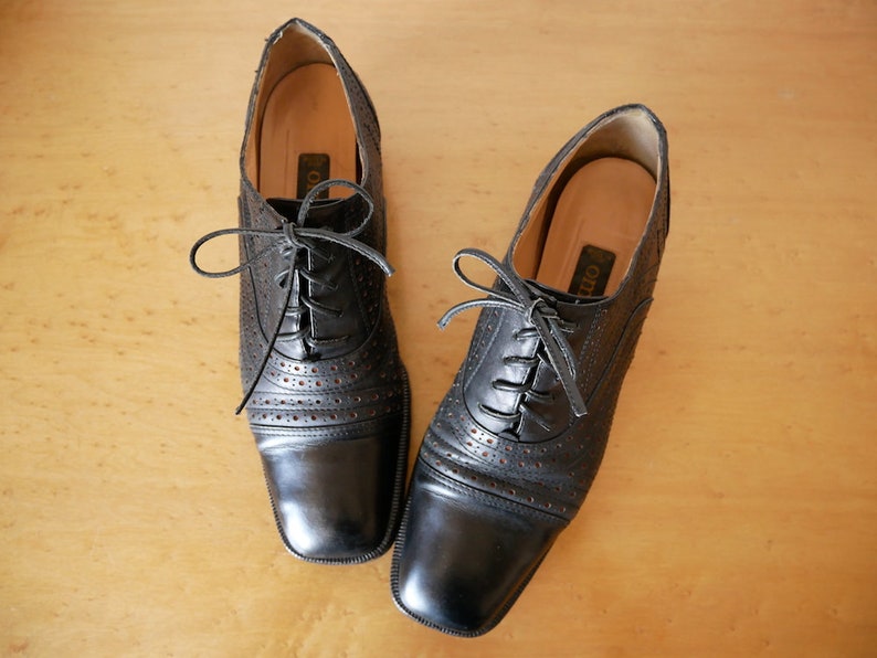May include: A pair of black leather lace-up shoes with a perforated design. The shoes have a square toe and a low heel.
