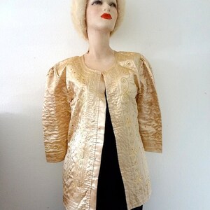 Peut inclure: Veste dor&eacute;e vintage avec broderies et ornements complexes. La veste a un col rond, des manches trois-quarts et une finition satin&eacute;e. La veste est pr&eacute;sent&eacute;e sur un mannequin portant un chapeau cr&egrave;me.