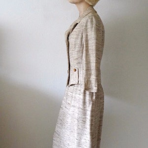 K&ouml;nnte beinhalten: Ein beiger Tweed-Rock-Anzug mit einer taillierten Jacke und einem geraden Rock. Die Jacke hat einen Knopfverschluss und eine Tasche auf der linken Seite. Der Rock hat eine hohe Taille und einen geraden Saum.