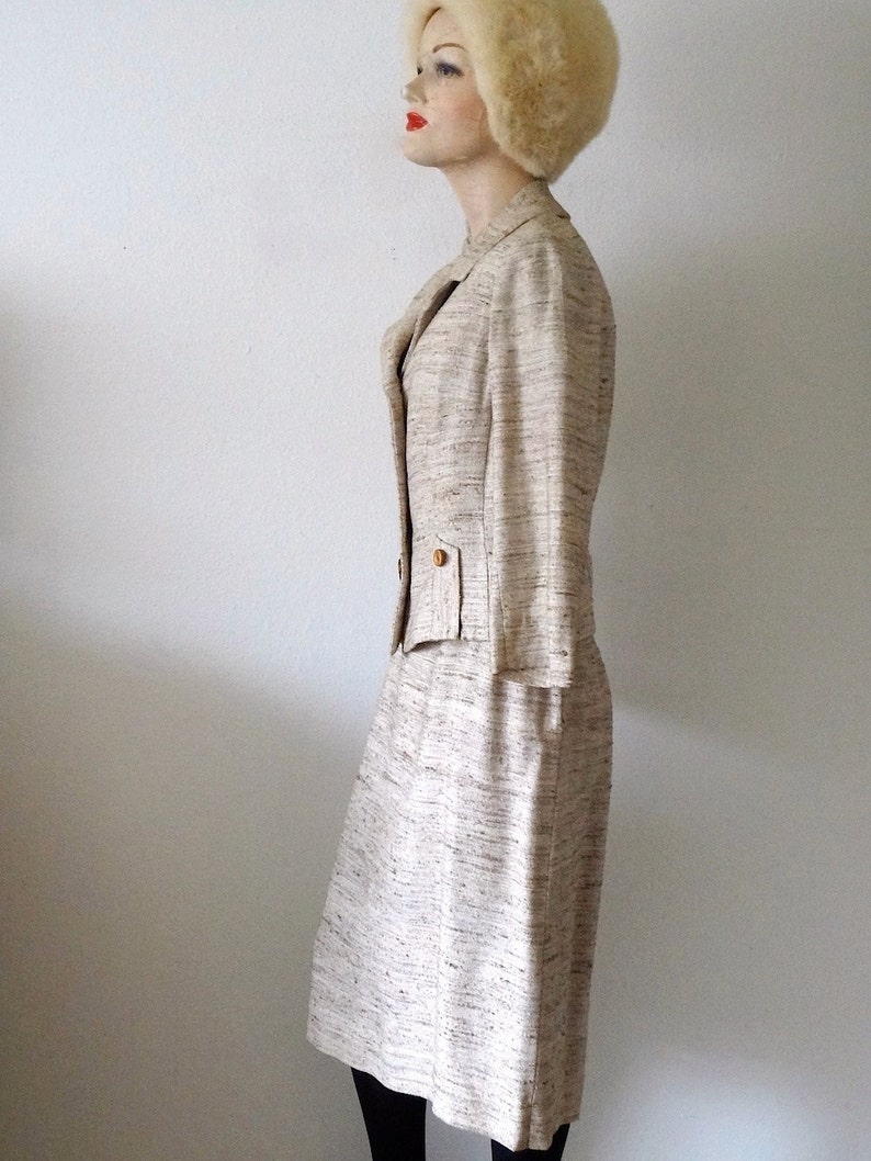 K&ouml;nnte beinhalten: Ein beiger Tweed-Rock-Anzug mit einer taillierten Jacke und einem geraden Rock. Die Jacke hat einen Knopfverschluss und eine Tasche auf der linken Seite. Der Rock hat eine hohe Taille und einen geraden Saum.