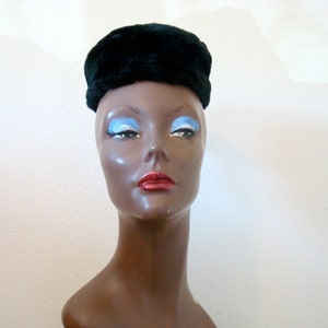 May include: A black faux fur pillbox hat. The hat is worn on a mannequin with brown skin and blue eyeshadow.