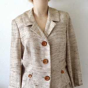K&ouml;nnte beinhalten: Ein beigefarbener Blazer mit braunem und wei&szlig;em Streifenmuster und Holzkn&ouml;pfen. Der Blazer hat einen Kragen und Taschen.
