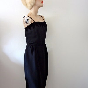 May include: A black sleeveless dress with a velvet trim. The dress is fitted and has a straight skirt. The model is wearing a white fur hat.