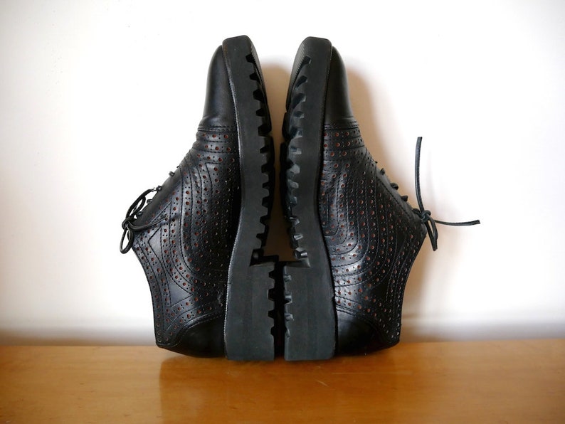 May include: A pair of black leather shoes with a lace-up closure and a textured sole. The shoes have a perforated design and are sitting on a wooden surface.
