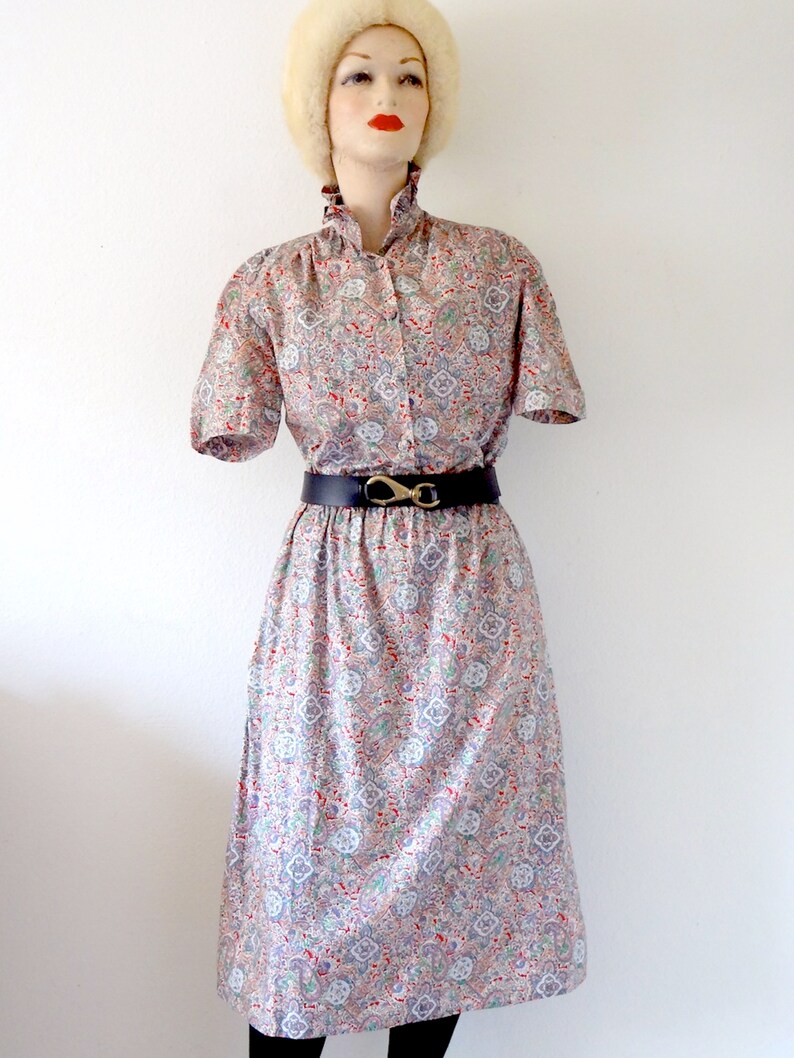 May include: A vintage dress with a paisley print in shades of pink, blue, and green. The dress has a short sleeve, a collared neckline, and a black belt with a gold buckle.