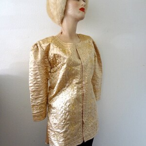Peut inclure: Veste dor&eacute;e &agrave; manches longues avec des motifs floraux et tourbillonnants brod&eacute;s. La veste pr&eacute;sente une encolure ronde et est orn&eacute;e de petits ornements dor&eacute;s. Un chapeau de fourrure assorti compl&egrave;te l'ensemble. La veste est pr&eacute;sent&eacute;e sur un mannequin.