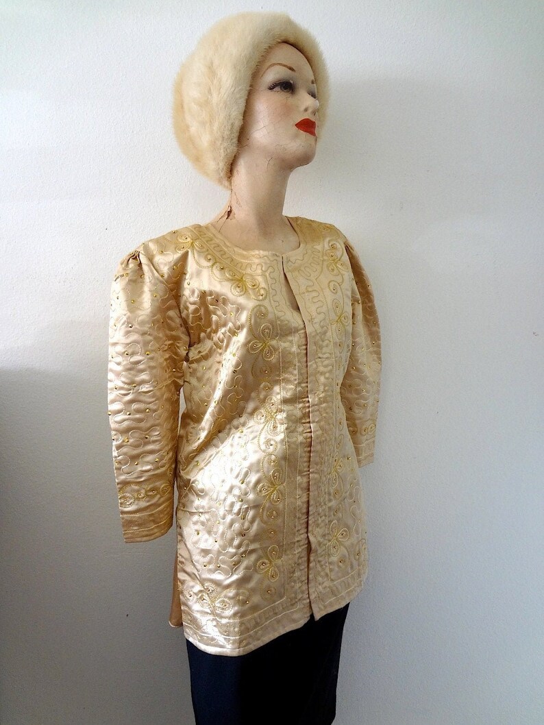 Peut inclure: Veste dor&eacute;e &agrave; manches longues avec des motifs floraux et tourbillonnants brod&eacute;s. La veste pr&eacute;sente une encolure ronde et est orn&eacute;e de petits ornements dor&eacute;s. Un chapeau de fourrure assorti compl&egrave;te l'ensemble. La veste est pr&eacute;sent&eacute;e sur un mannequin.