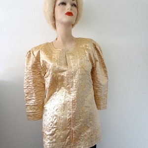 Peut inclure: Veste vintage de couleur or clair, orn&eacute;e de broderies florales et de motifs tourbillonnants. La veste pr&eacute;sente des manches trois-quarts et est orn&eacute;e de petits ornements scintillants. Le v&ecirc;tement est pr&eacute;sent&eacute; sur un mannequin, mettant en valeur son design et sa texture.