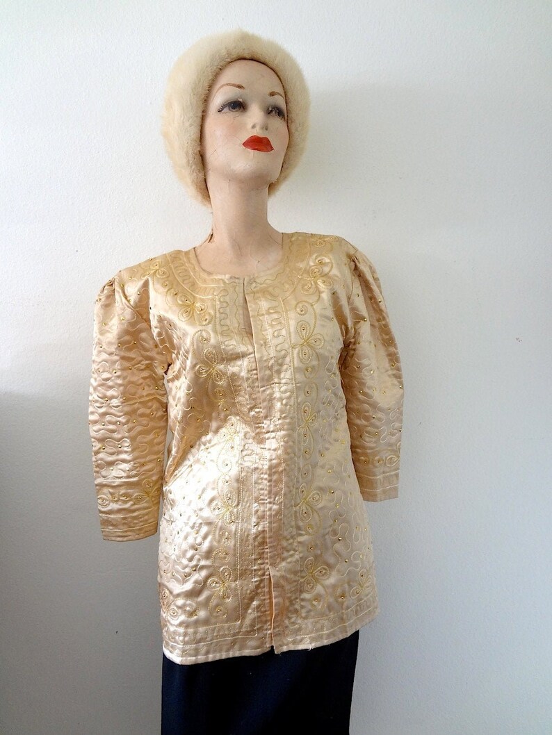 Peut inclure: Veste vintage de couleur or clair, orn&eacute;e de broderies florales et de motifs tourbillonnants. La veste pr&eacute;sente des manches trois-quarts et est orn&eacute;e de petits ornements scintillants. Le v&ecirc;tement est pr&eacute;sent&eacute; sur un mannequin, mettant en valeur son design et sa texture.
