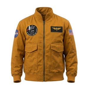 May include: Mustard-colored bomber jacket with a zip-up front, two front pockets, and a ribbed collar and cuffs. Features NASA and US flag patches, plus a circular patch with text. A rectangular patch with a winged emblem is also visible.