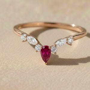 Op de afbeelding: Roségouden ring met een peervormige robijn als middensteen en kleine witte accentstenen. De ring heeft een gebogen ontwerp, met de robijn aan de onderkant van de boog. Op een neutrale achtergrond.