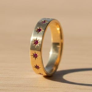 May include: A gold ring with a series of red star-shaped gemstones. The ring is set against a light brown wooden surface, with a shadow cast by the ring. The ring is a band style, with the gemstones evenly spaced around the band.