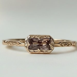 May include: A gold ring featuring a rectangular, brown gemstone within an ornate, rectangular setting. The band is decorated with swirling patterns. The ring is presented on a white surface, highlighting its intricate design and warm tones.