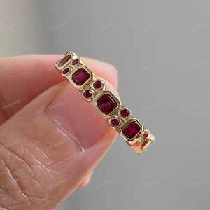 May include: A gold ring featuring alternating square and round red gemstones. The ring has a repeating pattern of square gemstones with round gemstones in between. The ring is held in a hand.