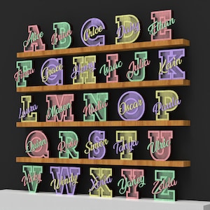 May include: Wooden shelves displaying colorful, 3D letter cutouts, each with a name. The letters are in various pastel colors, with names in cursive script. The shelves are brown, set against a dark gray background.