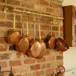 Peut inclure: Une cuisine avec des ustensiles de cuisine en cuivre suspendus à une barre en laiton sur un mur de briques. Une bouilloire en cuivre est sur la cuisinière, et un chou repose sur un plan de travail en bois. Un moulin à café vintage et un livre de cuisine ouvert sont également visibles.