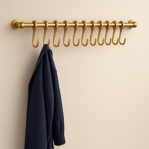 Könnte beinhalten: Ein goldfarbener Metall-Kleiderständer mit zehn gebogenen Haken. Eine marineblaue Jacke hängt an einem der Haken. Der Ständer ist an einer hellbeigen Wand befestigt.
