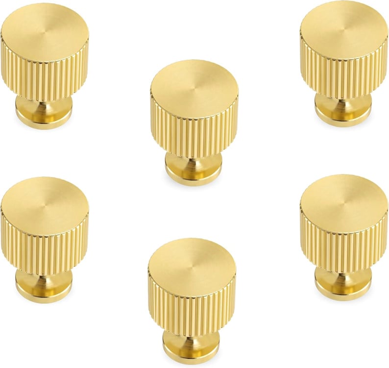 Pode incluir: Seis puxadores dourados para arm&aacute;rios. Cada puxador tem formato cil&iacute;ndrico com borda canelada e topo plano e circular. Os puxadores s&atilde;o feitos de um material met&aacute;lico brilhante.