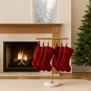 Puede incluir: Seis calcetines navideños de terciopelo rojo cuelgan de un soporte de metal dorado con base de mármol blanco. Una chimenea con fuego y un árbol de Navidad se encuentran en el fondo.
