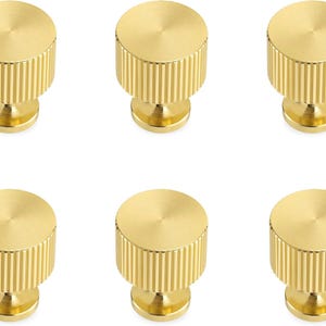 Pode incluir: Seis puxadores dourados para arm&aacute;rios. Cada puxador tem formato cil&iacute;ndrico com textura canelada nas laterais e um topo liso e circular. Os puxadores s&atilde;o montados em uma pequena base arredondada.
