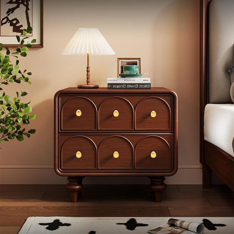 Pode incluir: Uma mesa de cabeceira de madeira castanha escura com seis gavetas, cada uma com um puxador dourado. Um abajur branco est&aacute; no topo, com uma pequena imagem emoldurada e livros. A mesa tem um design curvo e assenta em pernas curtas e arredondadas. A mesa tem cerca de 60 cm de largura.