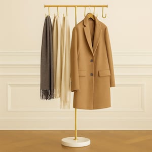 Puede incluir: Un perchero de metal dorado con una base circular blanca. Un abrigo color canela cuelga de una percha de madera, con dos bufandas beige y gris. El perchero tiene múltiples ganchos.