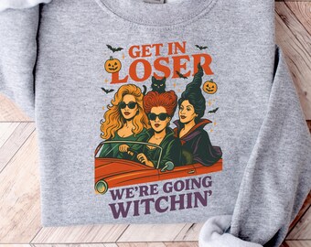 May include: A heather gray sweatshirt with the text "GET IN LOSER WE'RE GOING WITCHIN'". The graphic features three women in a car with a black cat, bats, and a pumpkin. The sweatshirt is folded on a wooden surface.