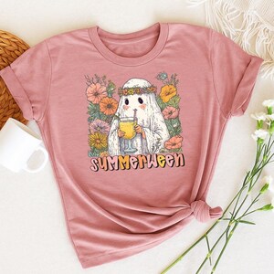 May include: A dusty pink t-shirt with a graphic of a ghost wearing a flower crown and holding a drink. The ghost is surrounded by flowers, with the word "Summerween" in a retro font below the image. The t-shirt is knotted at the bottom.