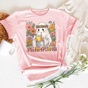 May include: A pale pink t-shirt with a graphic of a ghost wearing a floral crown and holding a drink. The word "SUMMERWEEN" is printed in a retro font below the ghost. The design is surrounded by colourful flowers.