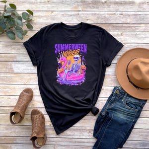 Może przedstawiać: Czarny t-shirt z kolorową grafiką przedstawiającą szkielet na dmuchanym flamingu, otoczony dyniami, z napisem "Summerween Vibes". Koszulka jest połączona z niebieskimi dżinsami i brązowym kapeluszem.