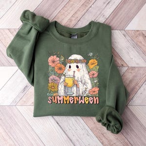 May include: Green sweatshirt with a ghost graphic wearing a floral crown and holding a drink. The design includes colourful flowers and the word "SUMMERWEEN". The sweatshirt has cuffed sleeves.