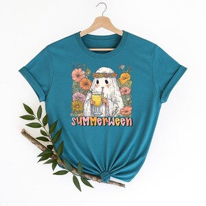 May include: Teal t-shirt with a cartoon ghost wearing a flower crown and holding a drink. The ghost is surrounded by colourful flowers, with the word "SUMMERWEEN" in a playful font below the image. The shirt is hanging on a wooden hanger.