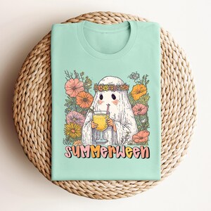 May include: Mint green t-shirt with a cartoon ghost wearing a flower crown and holding a drink. The ghost is surrounded by colourful flowers. The text "SUMMERWEEN" is printed in a retro font.