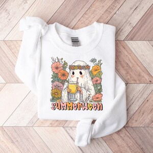 May include: White sweatshirt with a ghost character wearing a floral crown, holding a drink with a straw. The ghost is surrounded by colorful flowers. The word "SUMMERWEEN" is printed in a retro font.