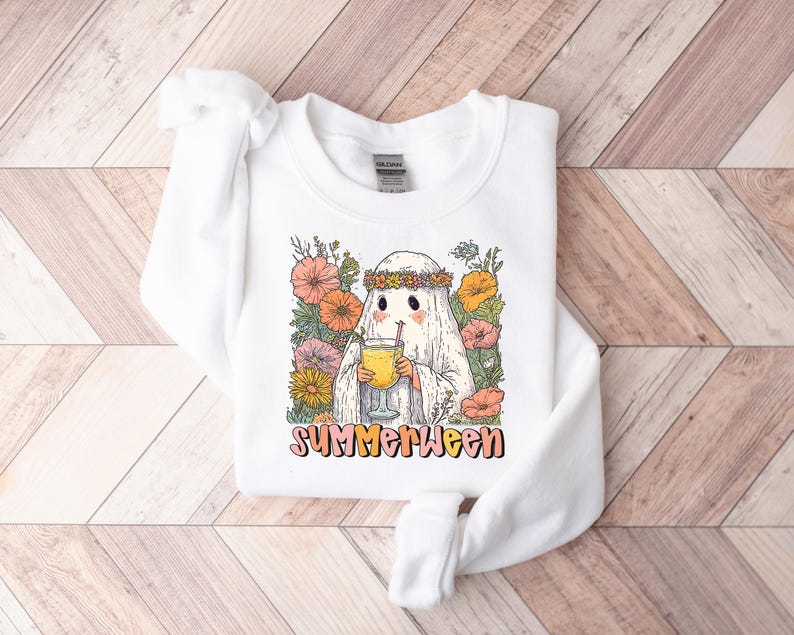 May include: White sweatshirt with a ghost character wearing a floral crown, holding a drink with a straw. The ghost is surrounded by colorful flowers. The word "SUMMERWEEN" is printed in a retro font.
