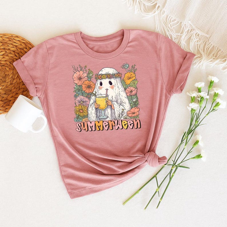 May include: A dusty pink t-shirt with a graphic of a ghost wearing a flower crown and holding a drink. The ghost is surrounded by flowers, with the word "Summerween" in a retro font below the image. The t-shirt is knotted at the bottom.
