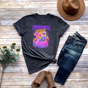 Może przedstawiać: Ciemnoszara koszulka z napisem "Summerween Vibes" i grafiką przedstawiającą szkielet na dmuchanym flamingu. Na zdjęciu znajdują się r&oacute;wnież niebieskie dżinsy, brązowy kapelusz i brązowe sandały.