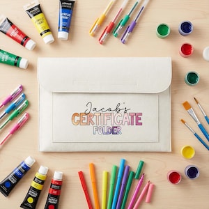 May include: A cream certificate folder with the text "Jacob's CERTIFICATE FOLDER" in colourful letters. The image also shows paint tubes, paintbrushes, pens, and small paint pots in various colours, suggesting an art or school theme.