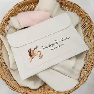 May include: A cream-colored baby announcement envelope with a stork illustration carrying a baby. The text "Baby Baker DUE JULY 2026" is printed on the front. The envelope rests in a wicker basket with soft, white baby blankets and a pink pillow.