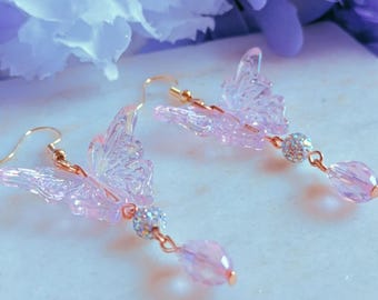 May include: A pair of dangle earrings featuring translucent, iridescent butterfly-shaped charms in a light purple hue. Each earring includes a small, round, silver-toned crystal-encrusted bead and a teardrop-shaped purple crystal suspended by gold-toned wire.