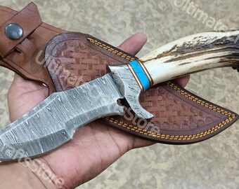 Peut inclure: Couteau de chasse en acier de Damas avec un manche en os et un accent turquoise, posé sur un étui en cuir marron. Le couteau a une lame texturée et un manche décoratif. L'étui est gaufré d'un motif.
