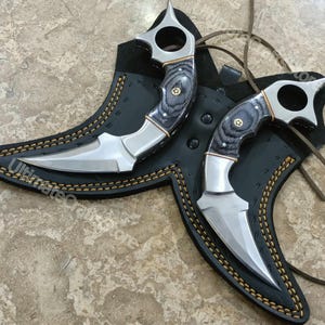 May include: Two curved knives with silver blades and gray and black wooden handles. The handles have a circular opening at the top. The knives are in black leather sheaths with gold stitching. The text "UltimateCraftStore" is visible.