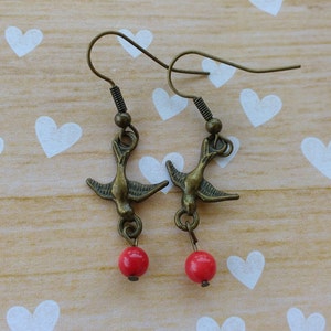 May include: A pair of bronze bird earrings with red bead accents. The earrings are on a wooden surface with a white heart pattern.