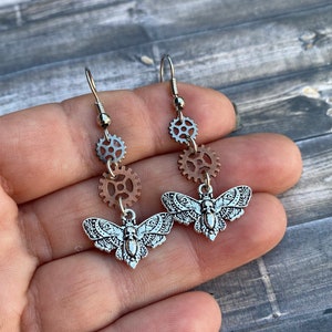 Op de afbeelding: Zilveren en koperen steampunk oorbellen met een gedetailleerde mot-hanger en drie tandwiel-hangers die aan elke oorbel hangen.